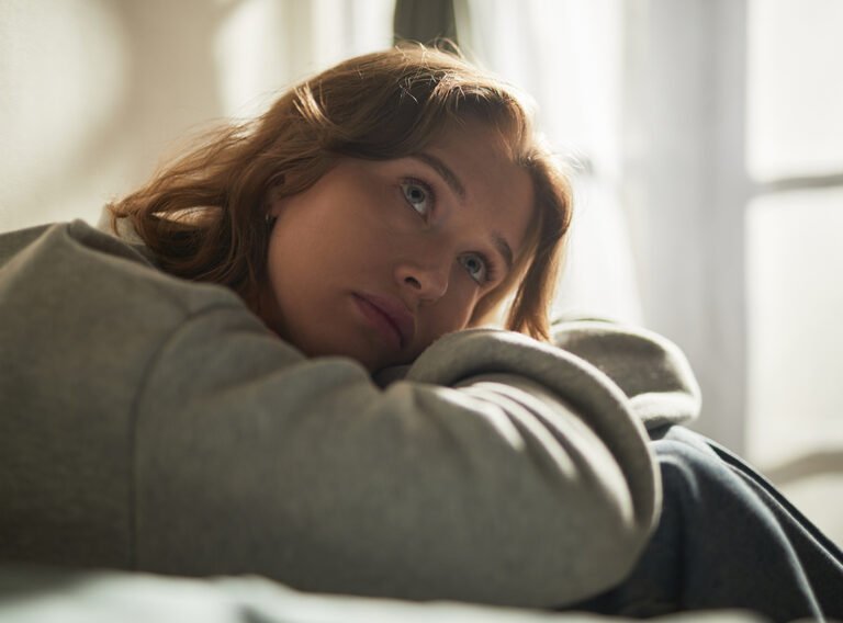 Caucasian young adult woman sitting indoors resting head on arms gazing thoughtfully into distance showing pensive facial expression conveying emotion of contemplation or sadness